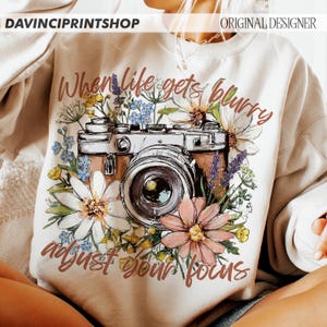 May include: Cream-colored sweatshirt with a floral and camera graphic. The design features a vintage camera surrounded by colorful flowers and the text "When life gets blurry, adjust your focus."