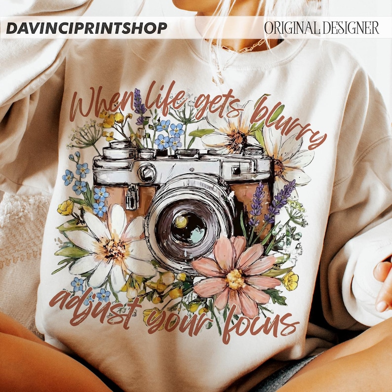 May include: Cream-colored sweatshirt with a floral and camera graphic. The design features a vintage camera surrounded by colorful flowers and the text "When life gets blurry, adjust your focus."