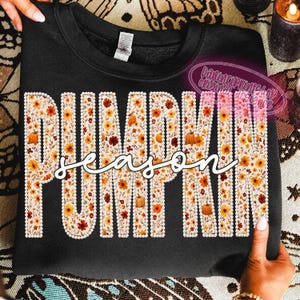 May include: A black crewneck sweatshirt featuring the word PUMPKIN in large block letters filled with a floral and pumpkin pattern, outlined in white faux pearls. The word season is written in white cursive script across the center.