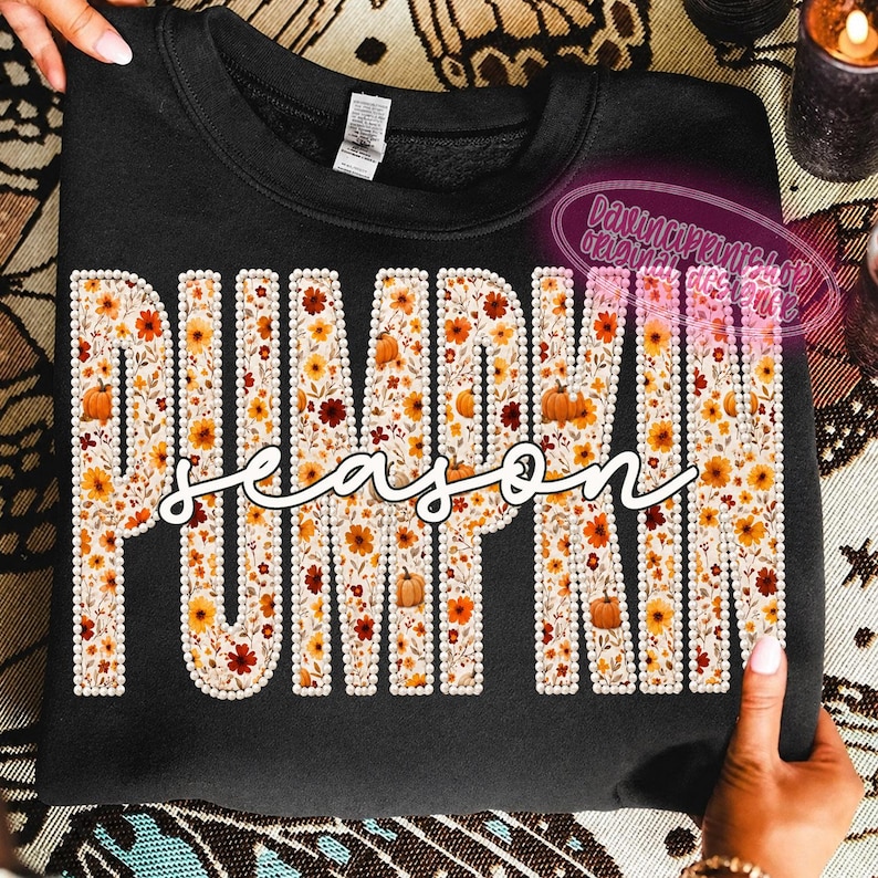May include: A black crewneck sweatshirt featuring the word PUMPKIN in large block letters filled with a floral and pumpkin pattern, outlined in white faux pearls. The word season is written in white cursive script across the center.