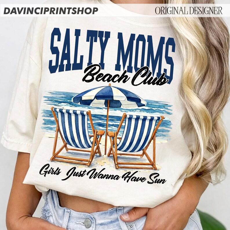 Op de afbeelding: Cr&egrave;mekleurig T-shirt met een strandthema. Het ontwerp toont de tekst "SALTY MOMS Beach Club" boven een illustratie van strandstoelen, een parasol en de tekst "Girls Just Wanna Have Sun."