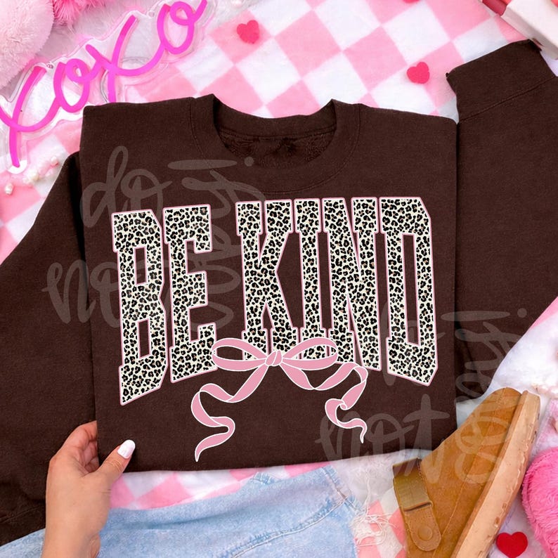 Op de afbeelding: Een bruine sweatshirt met de tekst "BE KIND" in een luipaardprint lettertype, met een roze strik eronder. Het kledingstuk wordt gepresenteerd op een roze en witte geruite achtergrond.