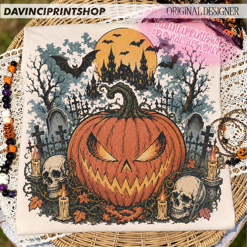 May include: A cream-coloured t-shirt featuring a graphic print of a large orange jack-o'-lantern with a jagged face, surrounded by two human skulls, lit candles, a dark castle silhouette, a full moon, bats, and a graveyard scene.