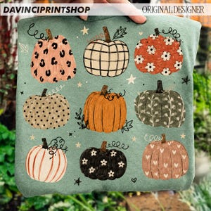 May include: A sage green sweatshirt featuring a graphic print of nine pumpkins in various patterns, including leopard print, plaid, floral, polka dot, and heart motifs, interspersed with small white stars and black vine illustrations.