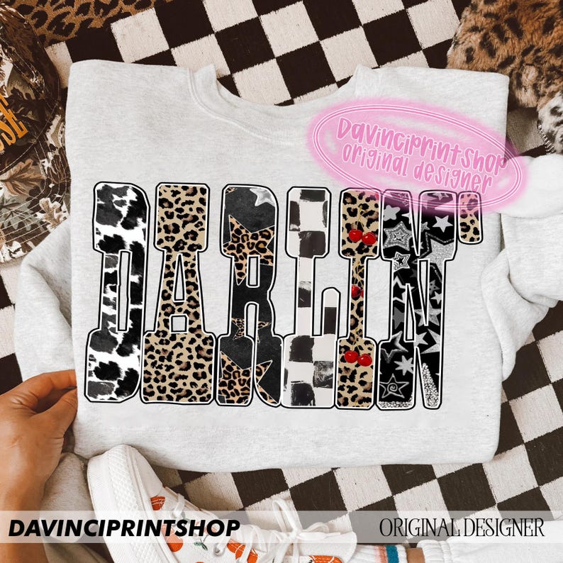 May include: A light gray sweatshirt with the word "DARLING" in large, bold letters. The letters are filled with various patterns, including cow print, leopard print, and stars. The text "DAVINCIPRINTSHOP ORIGINAL DESIGNER" is also visible.