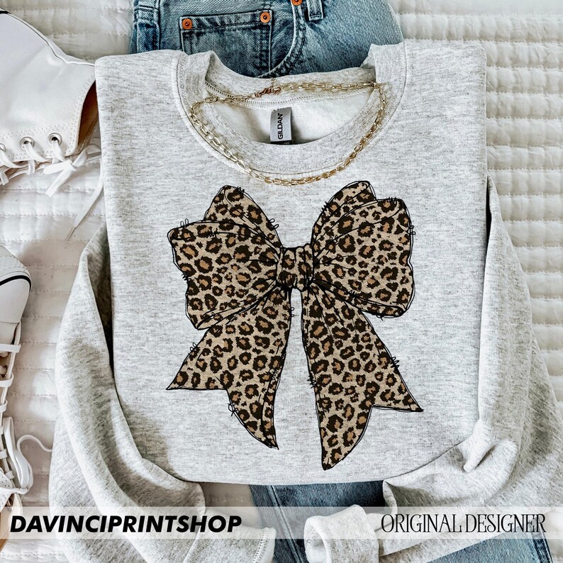 May include: A gray sweatshirt with a large leopard print bow design. The bow is centered on the sweatshirt and is a light brown color with black spots.