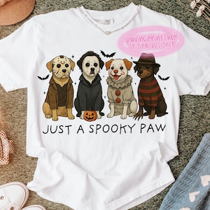 May include: White t-shirt featuring a graphic of four dogs dressed as horror movie characters. The dogs are wearing costumes from iconic films. The text "JUST A SPOOKY PAW" is printed below the dogs.