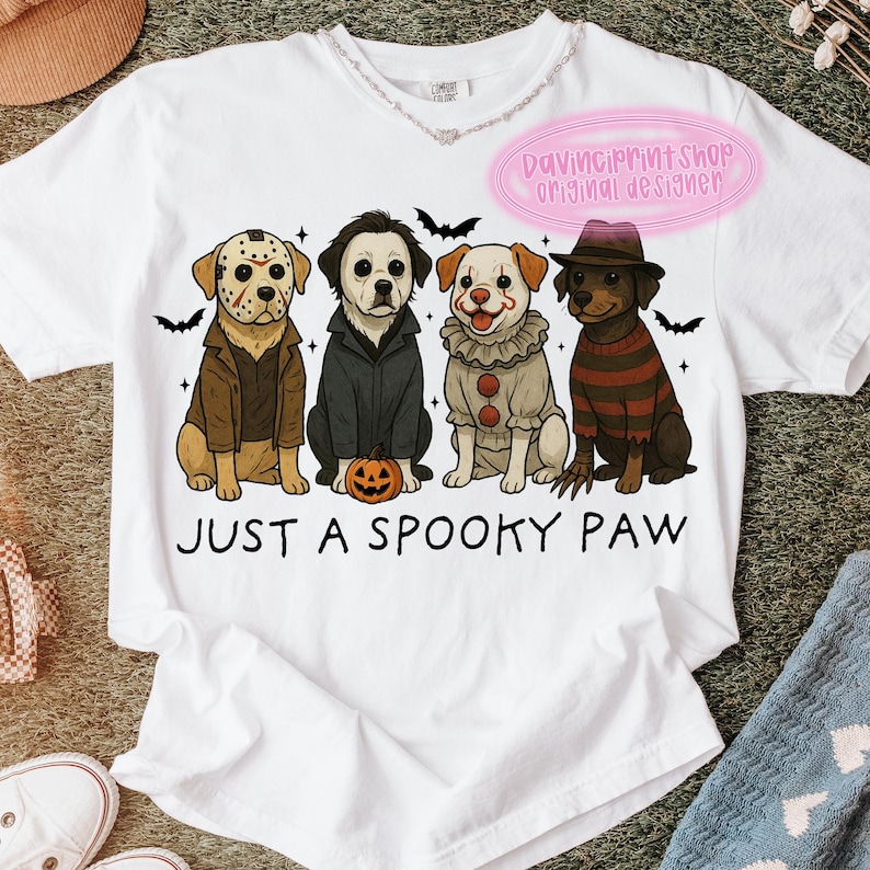 May include: White t-shirt featuring a graphic of four dogs dressed as horror movie characters. The dogs are wearing costumes from iconic films. The text "JUST A SPOOKY PAW" is printed below the dogs.