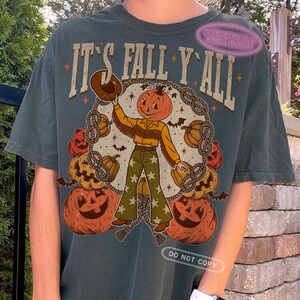 May include: Dark teal t-shirt with the text "IT'S FALL Y'ALL" above a pumpkin-headed cowboy graphic. The cowboy wears a brown hat, yellow shirt, and green pants with star patterns. Pumpkins and bats surround the cowboy.