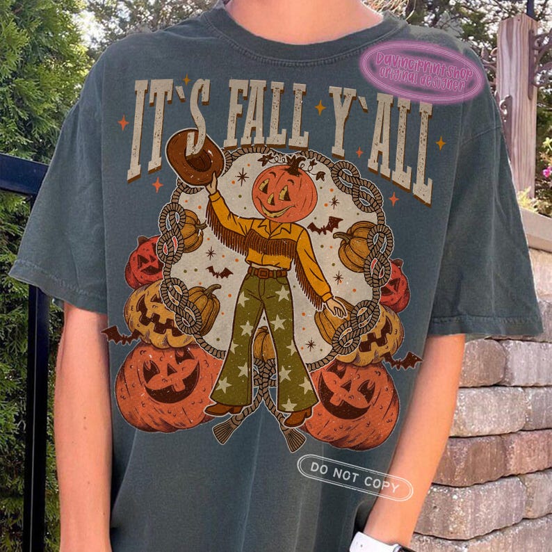 May include: Dark teal t-shirt with the text "IT'S FALL Y'ALL" above a pumpkin-headed cowboy graphic. The cowboy wears a brown hat, yellow shirt, and green pants with star patterns. Pumpkins and bats surround the cowboy.
