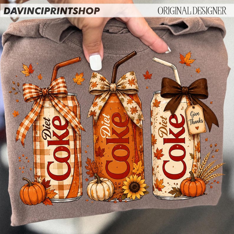 May include: A brown sweatshirt featuring a graphic of three Diet Coke cans with straws and bows. The cans are decorated with autumn themes including pumpkins, sunflowers, wheat, and fall leaves. One can has a tag that reads Give Thanks.