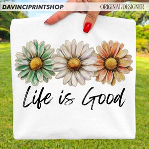 May include: White t-shirt with three daisy flowers in green, white, and orange, with yellow centres. The phrase "Life is Good" is written in black script below the flowers. The top left corner has the text "DAVINCIPRINTSHOP" and the top right corner has the text "ORIGINAL DESIGNER".