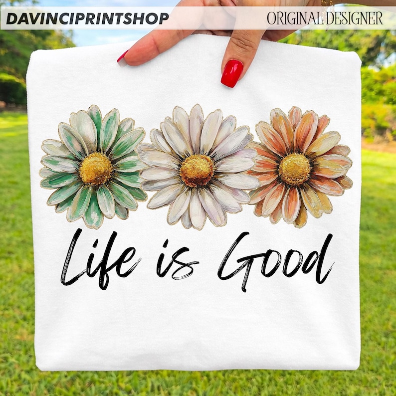 May include: White t-shirt with three daisy flowers in green, white, and orange, with yellow centres. The phrase "Life is Good" is written in black script below the flowers. The top left corner has the text "DAVINCIPRINTSHOP" and the top right corner has the text "ORIGINAL DESIGNER".