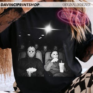 Peut inclure: Un t-shirt noir avec une impression graphique de personnages de films d'horreur assis dans une salle de cin&eacute;ma. Le motif montre des silhouettes portant des masques, dont un masque de hockey et un masque blanc, tenant du pop-corn. Texte DAVINCIPRINTSHOP.