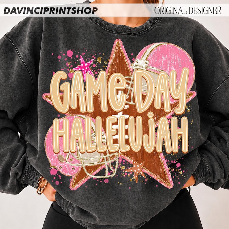 May include: A charcoal grey crewneck sweatshirt featuring a graphic print of a brown football, two pink football helmets, and pink stars. The text "GAME DAY HALLELUJAH" is printed in large, light tan letters across the center of the design.