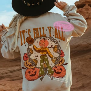 May include: Beige sweatshirt with a graphic featuring a pumpkin-headed cowboy holding a hat, surrounded by pumpkins and the text "It's Fall Y'all". The design includes stars, bats, and rope detailing, perfect for autumn.