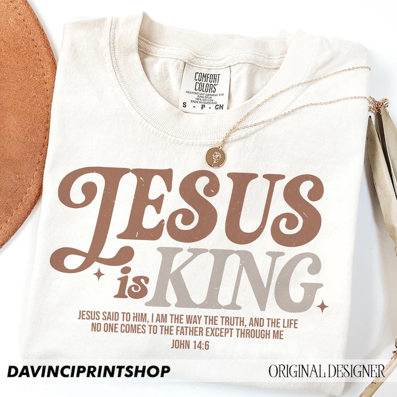 K&ouml;nnte beinhalten: Ein beiges T-Shirt mit dem Text "JESUS is KING." in braunen und grauen Buchstaben. Das Shirt enth&auml;lt auch einen Bibelvers aus Johannes 14,6: "Jesus sagte zu ihm: Ich bin der Weg, die Wahrheit und das Leben. Niemand kommt zum Vater au&szlig;er durch mich."