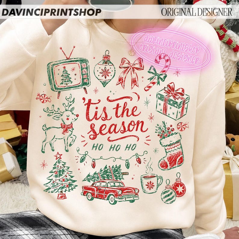 May include: A cream-colored sweatshirt featuring a red and green Christmas-themed graphic print. The design includes a vintage television, a reindeer, a car, a stocking, a gift, and the text 'tis the season' and 'HO HO HO' in red script.