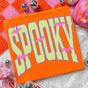May include: An orange t-shirt featuring the word SPOOKY printed in large, lime green and white vertical striped letters with a purple outline. Several small, bright pink bat silhouettes are scattered across the text design.