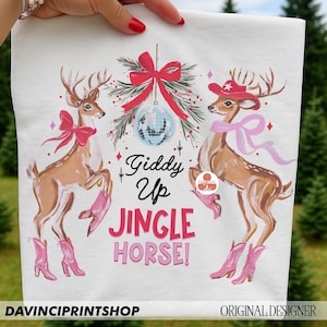 May include: White fabric with a festive design featuring two deer wearing pink cowboy boots and bows. The text "Giddy Up Jingle Horse!" is displayed in pink and black. A disco ball and red bow are also present.