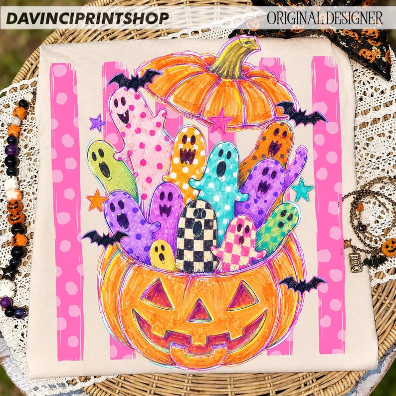 May include: A beige t-shirt featuring a graphic print of a large orange jack-o'-lantern filled with colorful, patterned ghosts. The design includes pink vertical stripes with white polka dots, small black bats, and various stars in the background.