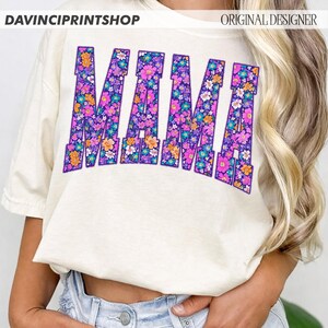 May include: Cream-colored t-shirt with the word "MAMA" in a floral design. The letters are purple with a variety of colorful flowers. The top of the shirt has the text "DAVINCIPRINTSHOP" and "ORIGINAL DESIGNER".
