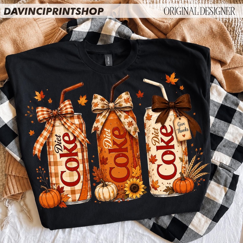 May include: A black t-shirt featuring a graphic of three Diet Coke cans decorated with autumn themes. Each can has a straw, a bow, and fall elements like pumpkins, sunflowers, and maple leaves. One can includes a tag that reads Give Thanks.