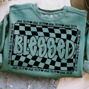 Op de afbeelding: Een turquoise sweatshirt met een zwart-wit geruit patroon en het woord "Blessed" in een vet, zwart lettertype. Het geruite patroon doet denken aan een racebaan. De tekst "John 20:29" wordt herhaald rond het geruite patroon.