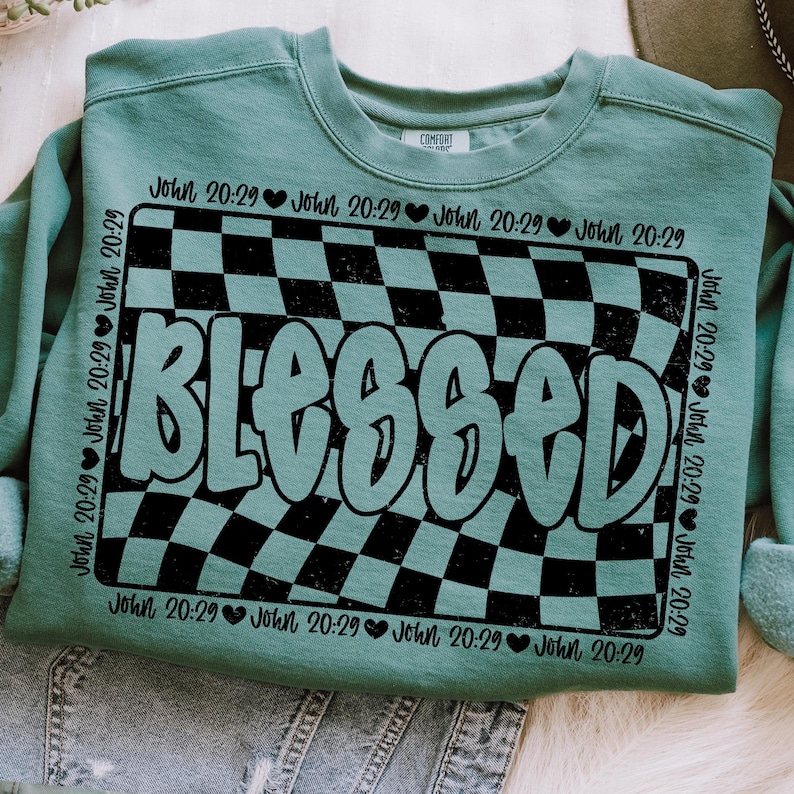 Op de afbeelding: Een turquoise sweatshirt met een zwart-wit geruit patroon en het woord "Blessed" in een vet, zwart lettertype. Het geruite patroon doet denken aan een racebaan. De tekst "John 20:29" wordt herhaald rond het geruite patroon.