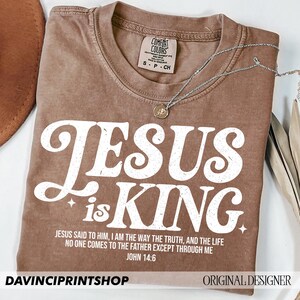 K&ouml;nnte beinhalten: Ein braunes T-Shirt mit einer wei&szlig;en Grafik, auf der in einer Retro-Schriftart "Jesus is King" steht. Das Shirt enth&auml;lt auch einen Bibelvers aus Johannes 14,6: "Jesus sagte zu ihm: Ich bin der Weg, die Wahrheit und das Leben. Niemand kommt zum Vater au&szlig;er durch mich."
