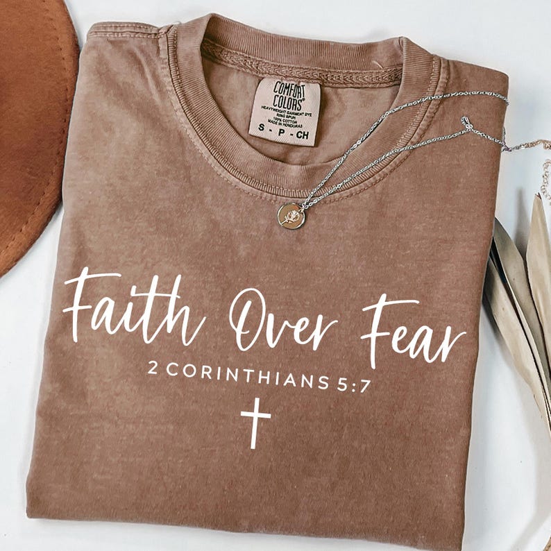 May include: A brown t-shirt with the text "Faith Over Fear 2 Corinthians 5:7" in white lettering. A white cross is below the text.