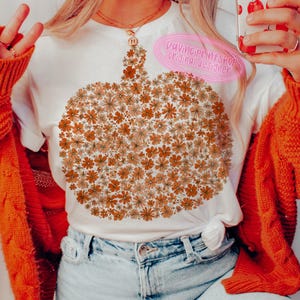 May include: White t-shirt featuring a pumpkin design made of orange and tan flowers. The shirt has a round neckline and short sleeves. The text "DaVinciPrintShop original designer" is on the upper right side of the shirt.