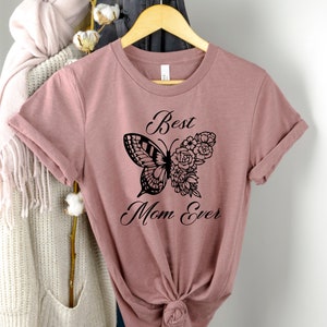 Pode incluir: Uma camiseta malva com um design de borboleta preta e flores. O texto "Best Mom Ever" est&aacute; impresso na camiseta.