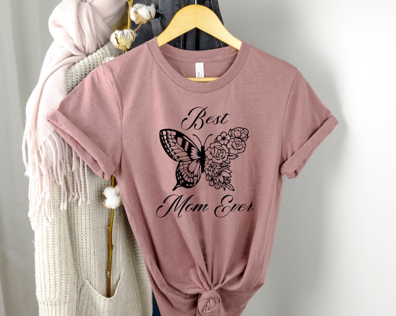 Pode incluir: Uma camiseta malva com um design de borboleta preta e flores. O texto "Best Mom Ever" est&aacute; impresso na camiseta.