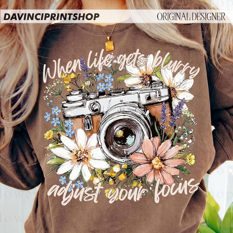 May include: Brown sweatshirt with a camera and floral design. The text reads "When life gets blurry adjust your focus." The camera is surrounded by white, pink, blue, and yellow flowers. The sweatshirt has a crew neck.