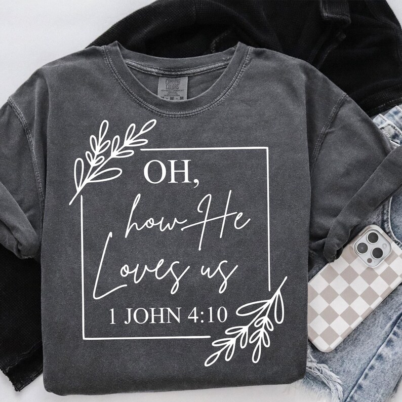 Pode incluir: Uma camiseta cinza escura com um design gr&aacute;fico branco que diz "OH, how He loves us 1 JOHN 4:10". O gr&aacute;fico &eacute; cercado por desenhos florais brancos.