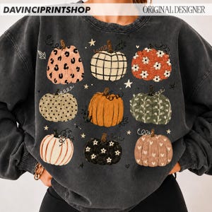 May include: A charcoal gray crewneck sweatshirt featuring a graphic print of nine pumpkins in various patterns, including leopard print, plaid, floral, polka dot, and heart motifs, interspersed with small white stars and vine-like flourishes.
