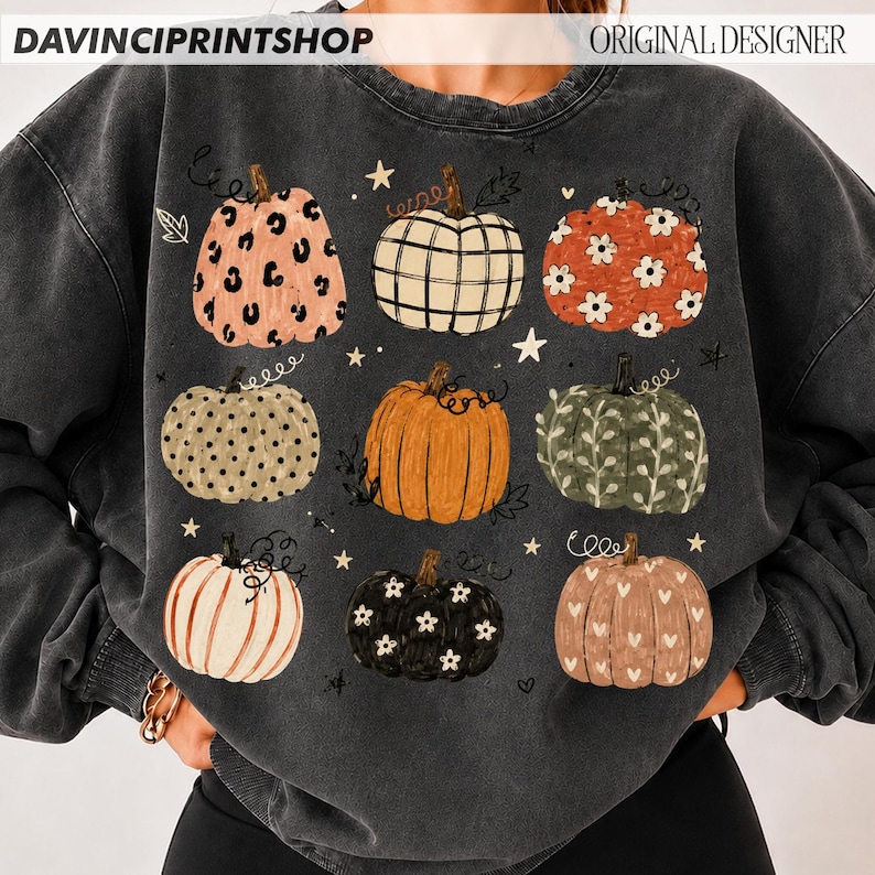 May include: A charcoal gray crewneck sweatshirt featuring a graphic print of nine pumpkins in various patterns, including leopard print, plaid, floral, polka dot, and heart motifs, interspersed with small white stars and vine-like flourishes.