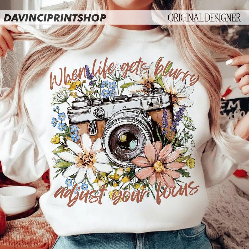 May include: White sweatshirt with a camera graphic surrounded by flowers. The text "When life gets blurry adjust your focus" is printed in brown script. The design includes a vintage camera, daisies, and other floral elements.