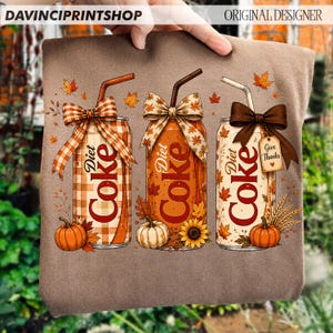 May include: A brown t-shirt featuring a graphic of three Diet Coke cans with straws, decorated with autumn-themed bows, falling leaves, pumpkins, and a sunflower. One can has a tag that reads Give Thanks, and the text Diet Coke is printed on each.
