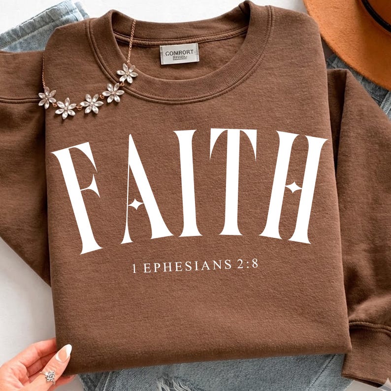 K&ouml;nnte beinhalten: Ein braunes Sweatshirt mit dem Wort "FAITH" in gro&szlig;en wei&szlig;en Buchstaben aufgedruckt. Unter dem Wort "FAITH" steht der Text "1 Ephesians 2:8". Eine goldene Halskette mit bl&uuml;tenf&ouml;rmigen Anh&auml;ngern liegt &uuml;ber dem Sweatshirt.