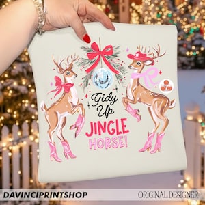 May include: A light beige cushion with a festive design featuring two deer wearing pink cowboy boots and accessories. The text "Gidy Up Jingle Horse!" is displayed in pink and black. The cushion is held by a person.