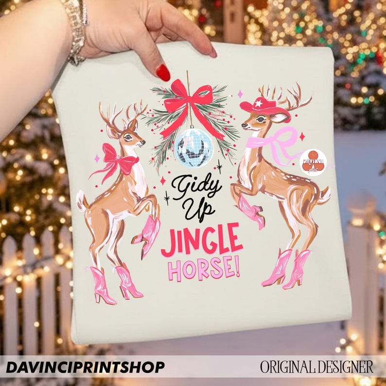 May include: A light beige cushion with a festive design featuring two deer wearing pink cowboy boots and accessories. The text "Gidy Up Jingle Horse!" is displayed in pink and black. The cushion is held by a person.
