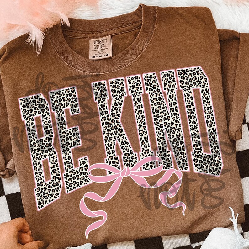 Op de afbeelding: Bruin t-shirt met de woorden "BE KIND" in een vetgedrukte luipaardprint lettertype, omlijnd in wit. Een roze strik staat onder de tekst. Het shirt heeft een vintage, versleten uitstraling.