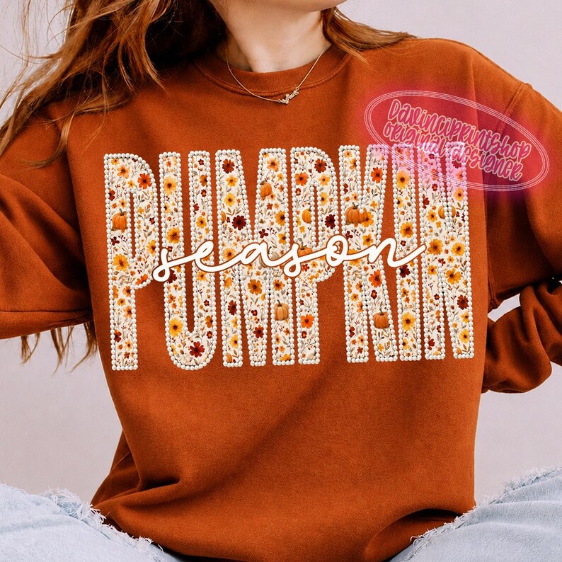 May include: A burnt orange crewneck sweatshirt featuring the word PUMPKIN in large block letters filled with a floral pattern of small orange, yellow, and red flowers and pumpkins, with the word season written in white cursive script across it.