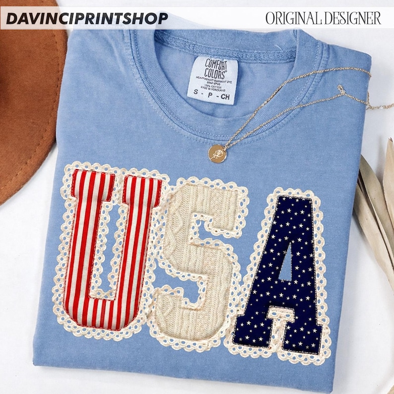 May include: A light blue t-shirt with the word "USA" appliqued on the front. The "U" is red with white stripes, the "S" is cable knit, and the "A" is navy blue with white stars. A gold necklace rests on the shirt.