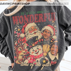 K&ouml;nnte beinhalten: Ein anthrazitfarbenes Sweatshirt mit Rundhalsausschnitt und einem Grafikdruck mit dem Text 'it's the most WONDERFUL time of the year'. Das Design zeigt Illustrationen vom Weihnachtsmann, einem Schneemann, einem Rentier und zwei Personen.