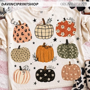 May include: A cream-colored t-shirt featuring a graphic print of nine pumpkins with various patterns including leopard print, plaid, floral, polka dots, and hearts, interspersed with small black stars and the text DAVINCIPRINTSHOP.