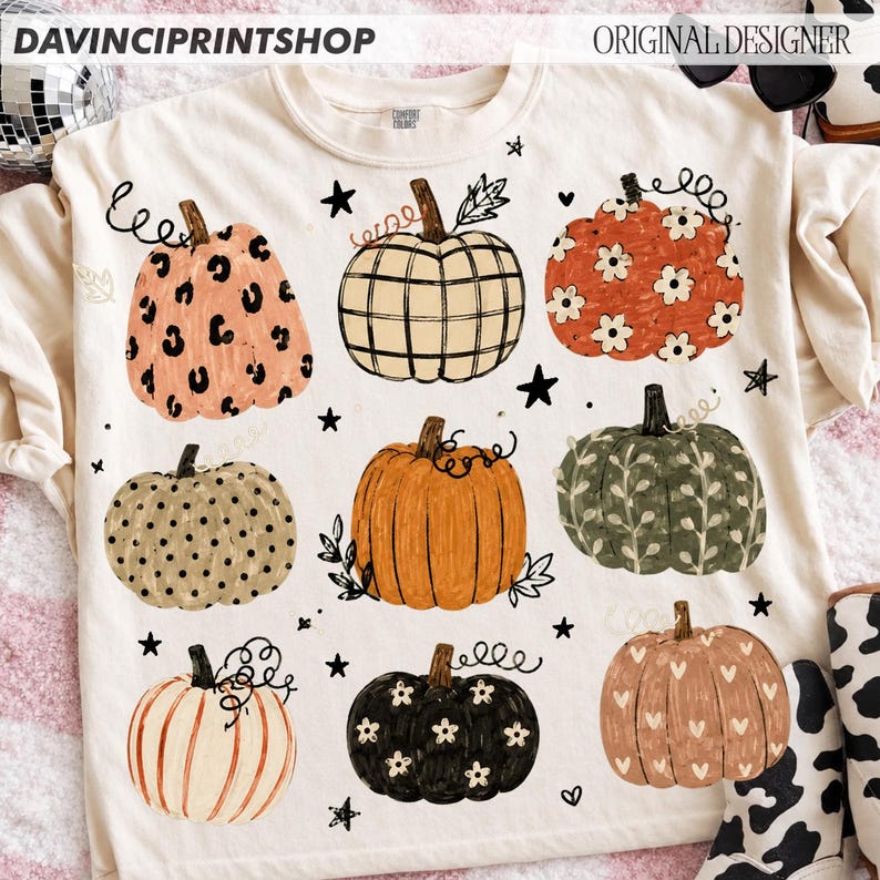 May include: A cream-colored t-shirt featuring a graphic print of nine pumpkins with various patterns including leopard print, plaid, floral, polka dots, and hearts, interspersed with small black stars and the text DAVINCIPRINTSHOP.