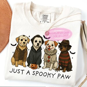 May include: Cream-colored t-shirt featuring four cartoon dogs dressed as horror movie characters. The phrase "JUST A SPOOKY PAW" is printed below the dogs. A gold necklace and a pink tag are also visible.